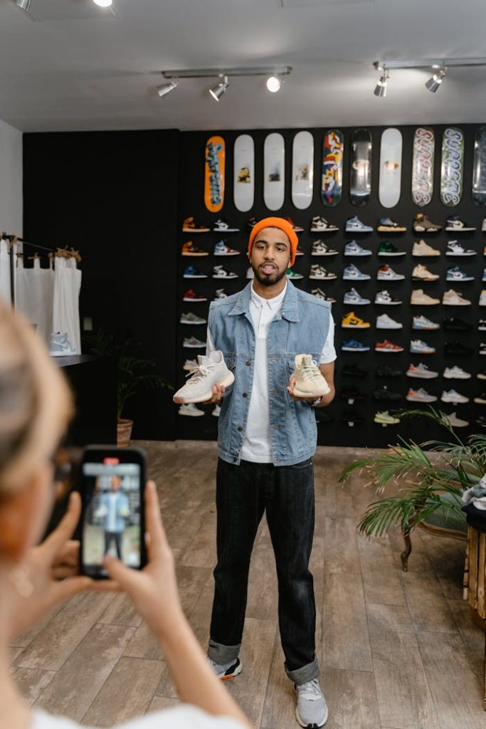 a-person-video-recording-a-man-in-denim-vest-holding-a-pair-of-rubber-shoes-7679677 A man demonstrates shoes for an online promotional video in a boutique shop setting.